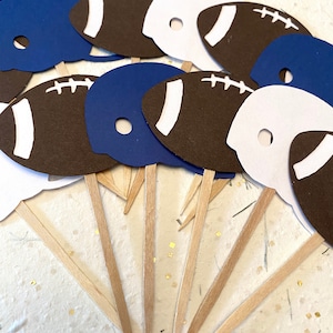 May include: A set of 10 football-themed cupcake toppers. The toppers are made of paper and feature a football helmet and a football. The helmets are blue and white, and the footballs are brown with white laces. The toppers are attached to wooden skewers.