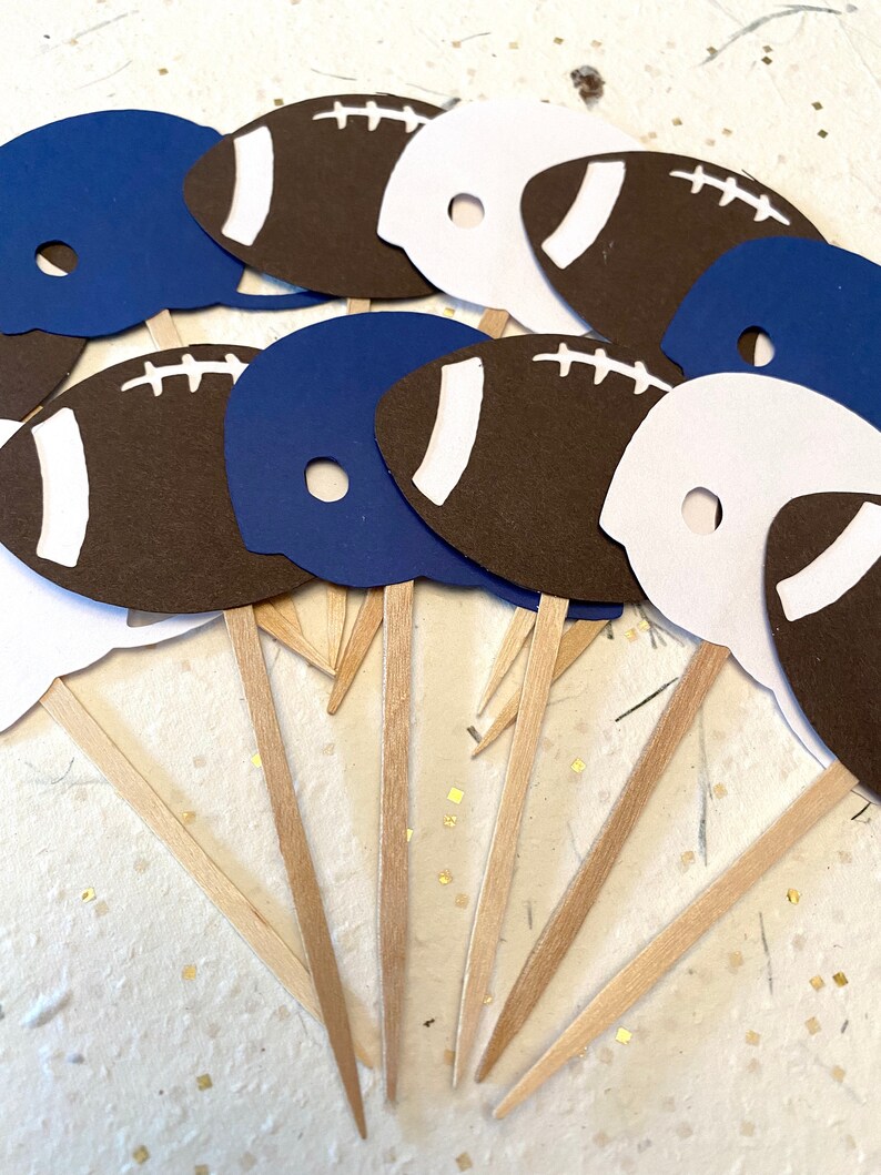 May include: A set of 10 football-themed cupcake toppers. The toppers are made of paper and feature a football helmet and a football. The helmets are blue and white, and the footballs are brown with white laces. The toppers are attached to wooden skewers.