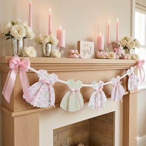 May include: A decorative banner featuring paper cutouts of dresses with floral and polka dot patterns in shades of pink and green, accented with pink ribbons. The banner is displayed on a wooden mantel with candles, flowers, and a sign reading It's a girl.
