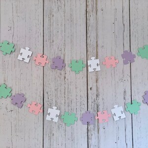 May include: A garland of pastel-colored puzzle pieces strung together with white thread. The puzzle pieces are in shades of pink, green, purple, and white.