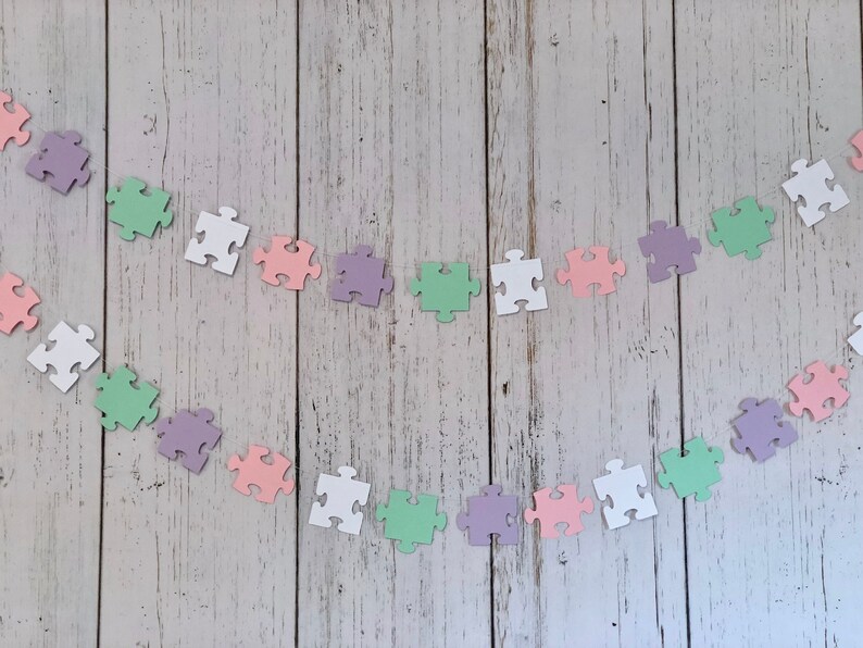 May include: A garland of pastel-colored puzzle pieces strung together with white thread. The puzzle pieces are in shades of pink, green, purple, and white.