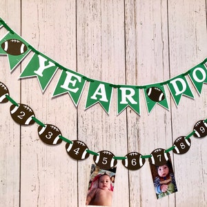 May include: Green and white banner with the text "1st Year Down" and football-shaped decorations with numbers 1 through 11. The banner is strung on a white wooden wall.