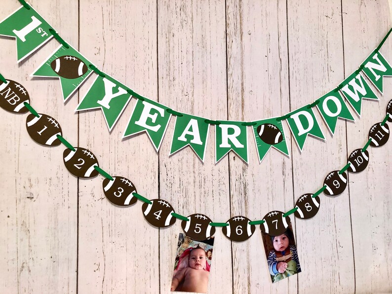 May include: Green and white banner with the text "1st Year Down" and football-shaped decorations with numbers 1 through 11. The banner is strung on a white wooden wall.