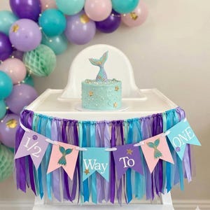 May include: A small round cake with light blue frosting, gold star accents, and a mermaid tail topper. A high chair banner features purple, pink, and blue pennants with the text 1/2 Way To ONE, decorated with mermaid tails, shells, and ribbon streamers.