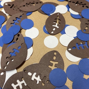 May include: Brown and white football confetti with blue and white polka dots. The confetti is scattered on a brown surface.