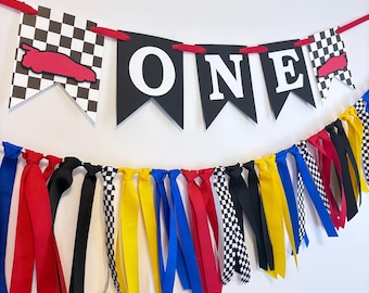 May include: A festive banner for a first birthday celebration. The banner spells "ONE" in white letters on black pennants. Checkered flags and red race cars accent the design. Colorful ribbons in red, blue, yellow, black, and checkered patterns hang below.