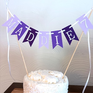 May include: A cake topper with a banner spelling "MADDIE 7" in silver letters on purple and lavender pennants. The banner is strung on wooden dowels and white ribbon, placed on top of a white frosted cake.