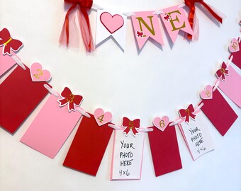 May include: A festive banner with red and pink elements, perfect for a first birthday celebration. The banner spells "ONE" with gold letters and includes heart-shaped accents and photo holders. Each photo holder is 4x6 inches.