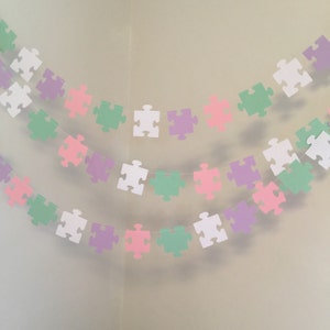 May include: A garland of pastel colored paper puzzle pieces strung together. The garland is made up of white, light green, light pink, and light purple puzzle pieces.