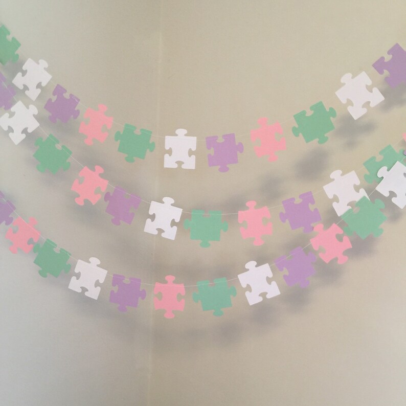 May include: A garland of pastel colored paper puzzle pieces strung together. The garland is made up of white, light green, light pink, and light purple puzzle pieces.