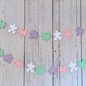 May include: A garland of pastel colored puzzle pieces strung together with white thread. The puzzle pieces are in shades of pink, purple, green, and white.