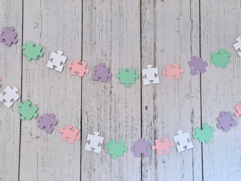 May include: A garland of pastel colored puzzle pieces strung together with white thread. The puzzle pieces are in shades of pink, purple, green, and white.