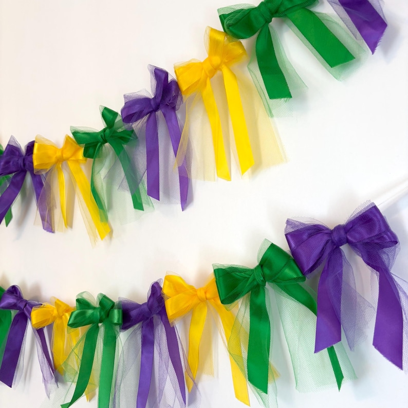 May include: A Mardi Gras party garland with purple, green, and gold bows made of tulle and ribbon. The garland is strung together with white string.