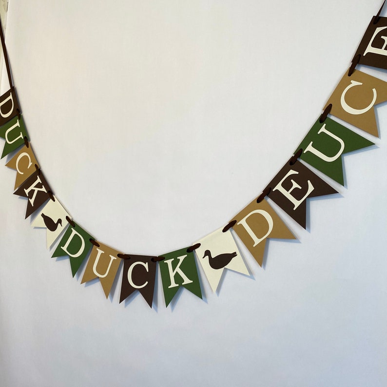 May include: A brown, green, and tan banner with the words "Duck, Duck, Deuce" in white letters. Each letter is on a separate triangular flag with a brown duck silhouette.