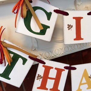 K&ouml;nnte beinhalten: Ein wei&szlig;es Banner mit dem Wort "GIVE THANKS" in gr&uuml;nen, orangefarbenen und gelben Buchstaben. Jeder Buchstabe befindet sich auf einem separaten wei&szlig;en Quadrat mit einem daran befestigten braunen Band. Die Quadrate sind mit kleinen braunen Bl&auml;ttern verziert.