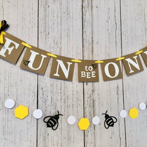 Puede incluir: Una pancarta de papel marr&oacute;n con las palabras "Fun to Bee One" en letras blancas. La pancarta est&aacute; decorada con peque&ntilde;as abejas negras y amarillas. Tambi&eacute;n se incluye una cadena separada de abejas negras, c&iacute;rculos blancos y hex&aacute;gonos amarillos.