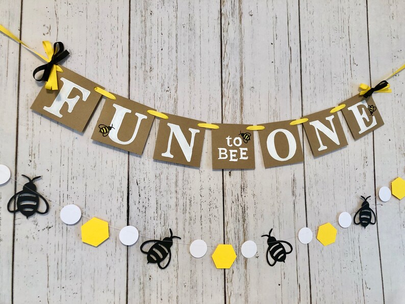 Puede incluir: Una pancarta de papel marr&oacute;n con las palabras "Fun to Bee One" en letras blancas. La pancarta est&aacute; decorada con peque&ntilde;as abejas negras y amarillas. Tambi&eacute;n se incluye una cadena separada de abejas negras, c&iacute;rculos blancos y hex&aacute;gonos amarillos.