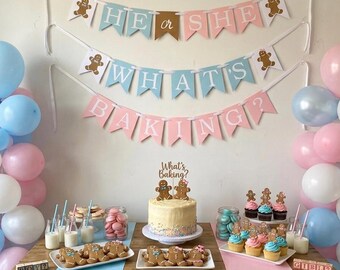 May include: A gender reveal party display featuring a three-tiered banner with the text HE OR SHE WHAT'S BAKING, a frosted cake with a cake topper, gingerbread cookies, cupcakes, macarons, and blue and pink balloons on a wooden table.
