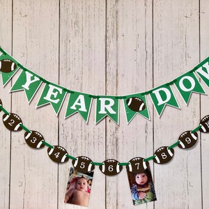 May include: Green and white banner with the words "1st Year Down" and football-shaped decorations. A second banner with football-shaped decorations numbered 1 through 11 is strung below the first banner. Photos of a baby are displayed in the football-shaped decorations.