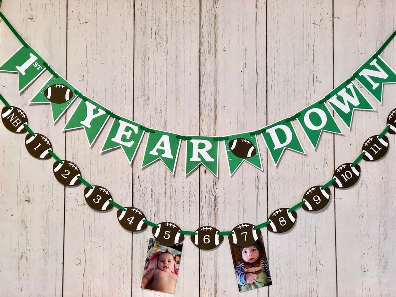 May include: Green and white banner with the words "1st Year Down" and football-shaped decorations. A second banner with football-shaped decorations numbered 1 through 11 is strung below the first banner. Photos of a baby are displayed in the football-shaped decorations.