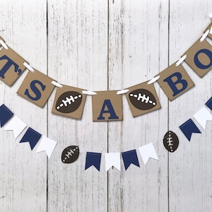May include: It's a Boy banner with brown and blue paper pennants. The pennants have the words "It's a Boy" and footballs printed on them. The banner is strung with white twine.