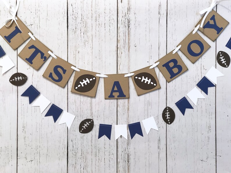 May include: It's a Boy banner with brown and blue paper pennants. The pennants have the words "It's a Boy" and footballs printed on them. The banner is strung with white twine.