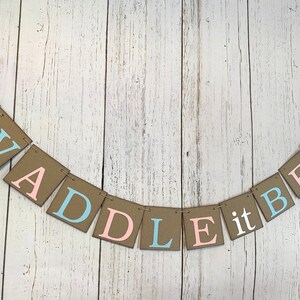 May include: A brown paper banner with the words "Waddle it be?" in pink and blue letters. The banner is decorated with a pink and blue duck silhouette on each end.