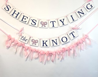 May include: White banner with blue text that reads "She's tying the knot" with pink bows and pink ribbon streamers.