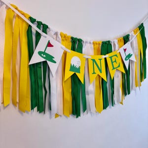 May include: A golf-themed birthday banner with white, yellow, and green fabric streamers. The banner has the word 