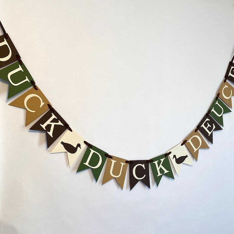 May include: A green, brown, and white banner with the word "DUCK DEUCE" in white letters. The banner has a duck silhouette on each side of the word "DUCK".