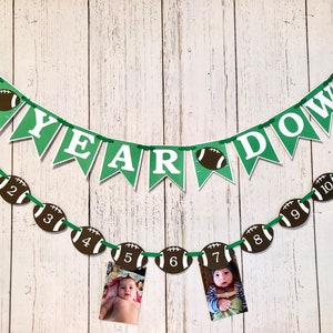 May include: Green and white banner with the text "1st Year Down" and a string of brown and white football-shaped decorations with numbers 1 through 12. Two photos are attached to the footballs.