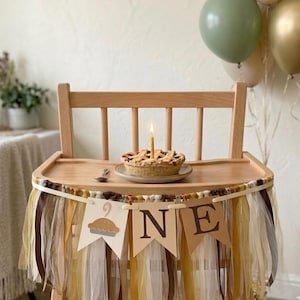 May include: A wooden high chair decorated with a fabric tassel banner in shades of brown, gold, and cream. The banner features pennants with a pie graphic and the letters O, N, E. A small pie with a single lit candle sits on the tray.