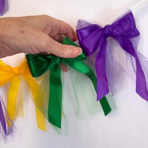 May include: A garland of tulle and satin ribbon bows in purple, green, and yellow. The bows are tied together with a white string.