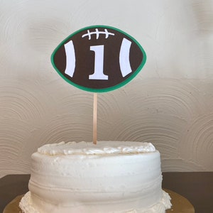 May include: A white cake with brown and green frosting decorated with a brown and white football with the number "1" on it.