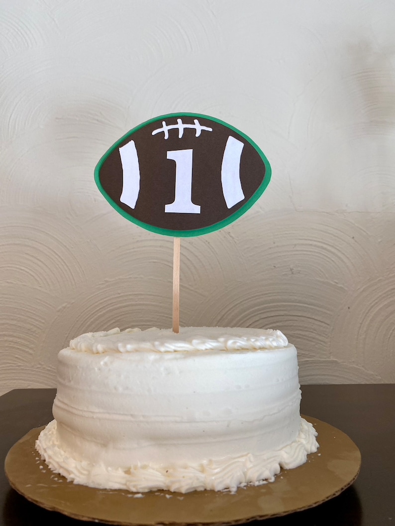 May include: A white cake with brown and green frosting decorated with a brown and white football with the number "1" on it.