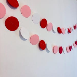 May include: Two strands of decorative paper garland with red, pink, and white circular cutouts. The garland is strung on a thin, clear thread and hangs against a white wall. The circles are various sizes.