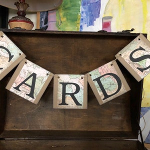 May include: A brown wooden box with a banner that reads "CARDS" in black letters. The banner is made of paper with a map design.