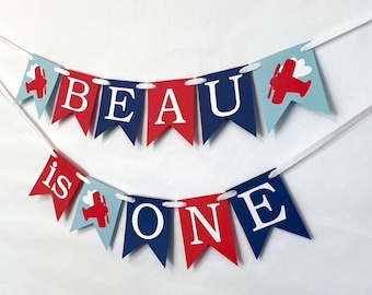 May include: A red, white, and blue banner with the words "BEAU is ONE" and red airplanes with white clouds.