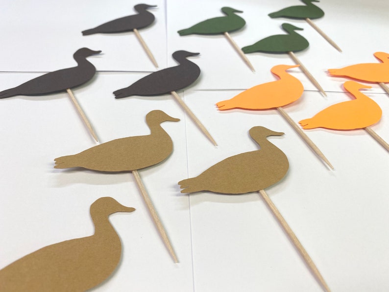 May include: Assortment of paper duck cake toppers in brown, green, and orange. The ducks are cut out and attached to wooden skewers.