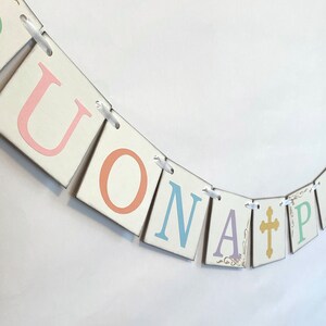 May include: A white banner with the words "BUONA PASQUA" in pastel colors. The letters are on individual squares with a white background and are connected by white ribbon. A gold cross is included in the banner.