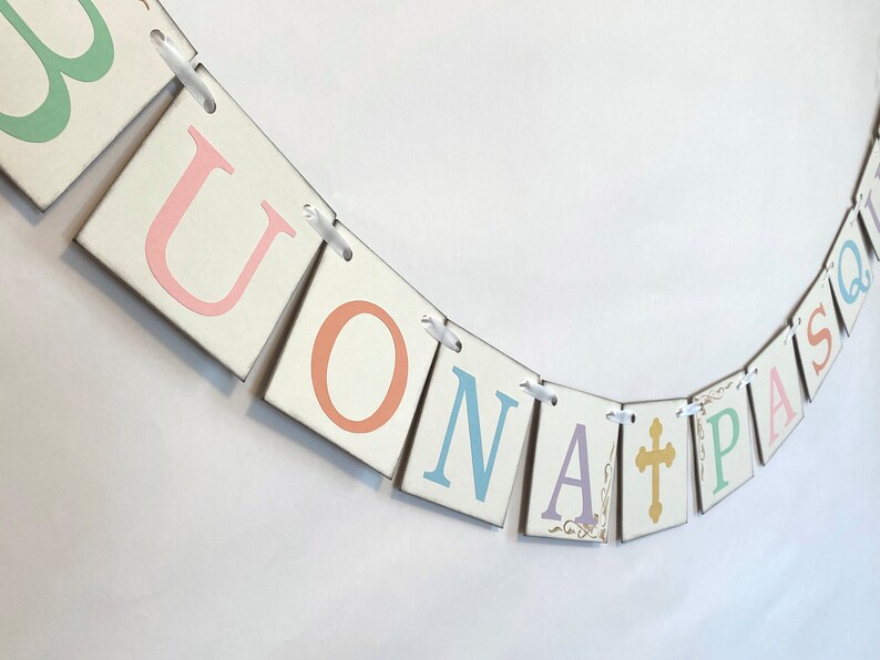May include: A white banner with the words "BUONA PASQUA" in pastel colors. The letters are on individual squares with a white background and are connected by white ribbon. A gold cross is included in the banner.