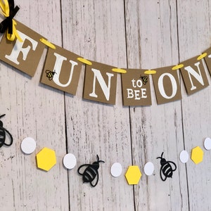 Puede incluir: Una pancarta de papel marr&oacute;n con el texto "Fun to Bee One" en letras blancas. La pancarta est&aacute; decorada con abejas amarillas y negras. Una cadena de c&iacute;rculos blancos, hex&aacute;gonos amarillos y abejas negras cuelga debajo de la pancarta.