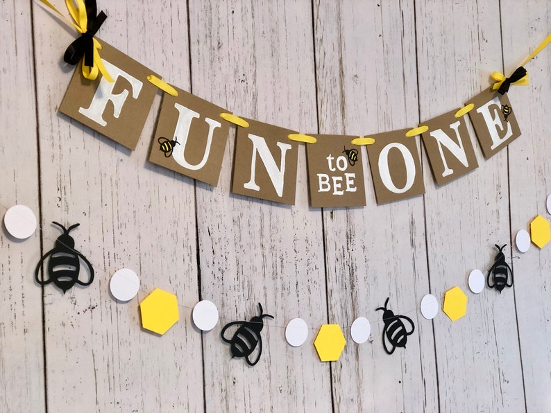 Puede incluir: Una pancarta de papel marr&oacute;n con el texto "Fun to Bee One" en letras blancas. La pancarta est&aacute; decorada con abejas amarillas y negras. Una cadena de c&iacute;rculos blancos, hex&aacute;gonos amarillos y abejas negras cuelga debajo de la pancarta.