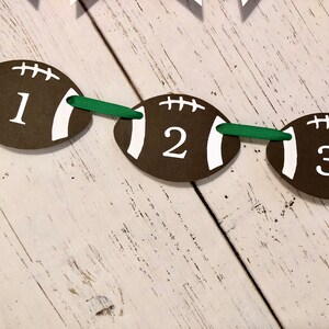 May include: A brown and white football-shaped banner with the numbers 1, 2, and 3 printed on it. The banner is strung together with green ribbon.