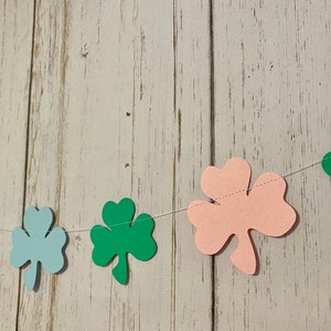Pu&ograve; includere: Una ghirlanda di trifogli verdi, blu e rosa infilata su uno sfondo di legno bianco. La ghirlanda &egrave; fatta di trifogli di carta.