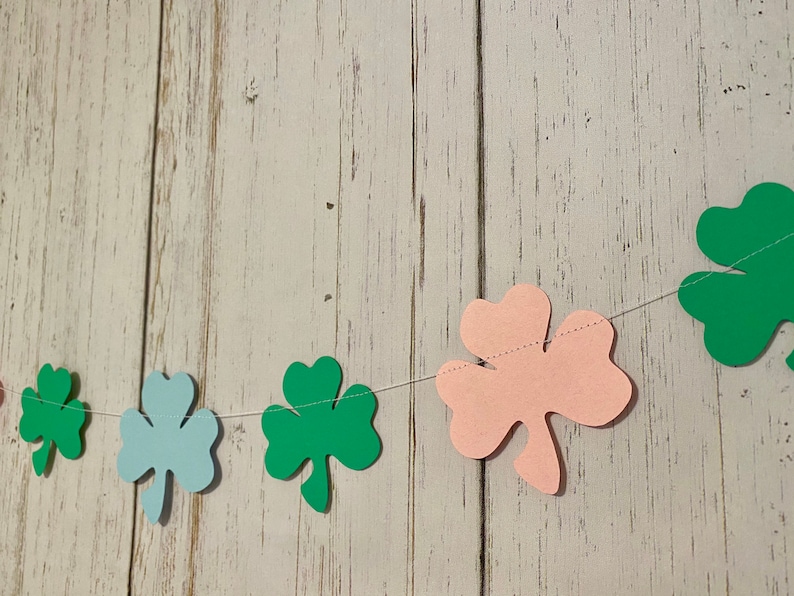 Pu&ograve; includere: Una ghirlanda di trifogli verdi, blu e rosa infilata su uno sfondo di legno bianco. La ghirlanda &egrave; fatta di trifogli di carta.