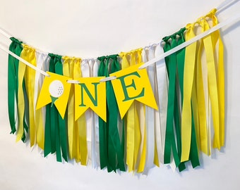 Op de afbeelding: Een gele en groene banner met het woord "ONE" en een golfbalafbeelding. De banner is versierd met groene, gele en witte linten.
