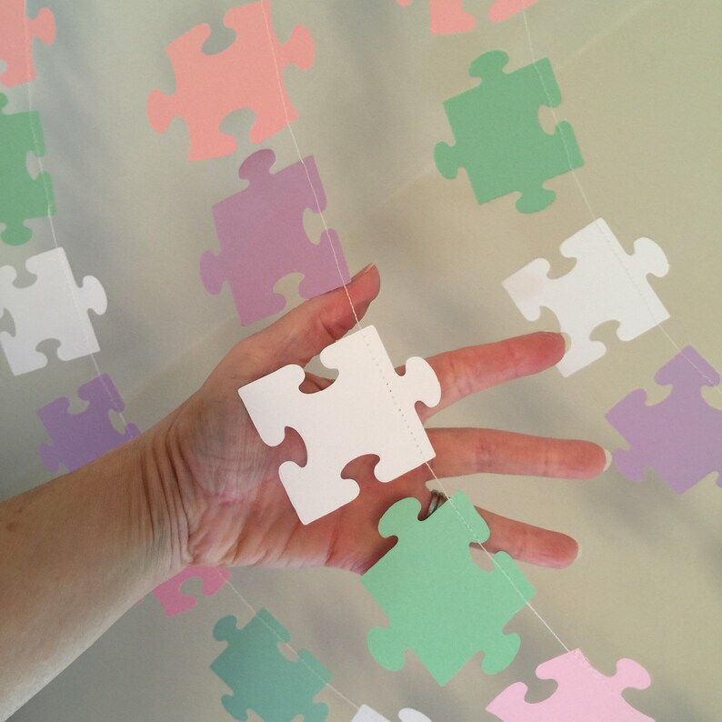 May include: A garland of pastel colored paper puzzle pieces strung together with white thread. The puzzle pieces are in pink, green, purple, and white.