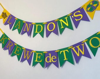 Puede incluir: Una pancarta festiva con las palabras "LANDON'S KREWE de TWO" en letras doradas brillantes. Las banderolas son moradas y verdes, con detalles amarillos y un diseño de corona y flor de lis. Ideal para una fiesta de cumpleaños.