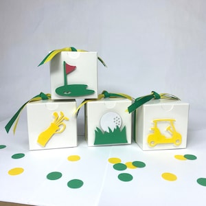 May include: Four white favor boxes with yellow and green ribbon. Each box has a different golf-themed design: a golf club, a golf ball, a golf cart, and a golf flag. The boxes are on a white surface with green and yellow confetti.